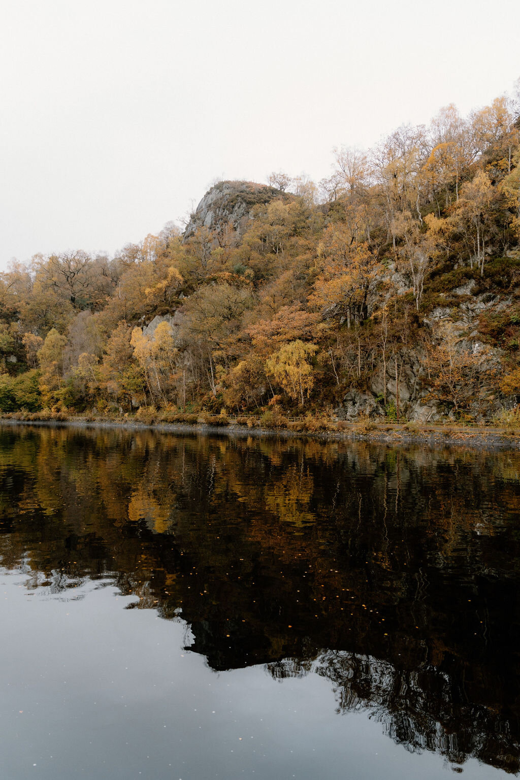 A rocky outcrop rises above autumn woodland reflected in a still loch near Wildwoodz, fallen leaves drifting on the water.