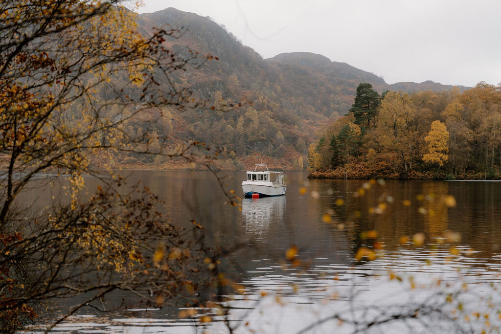A small tour boat glides across a calm loch near Wildwoodz, autumn branches framing the scene from both sides.