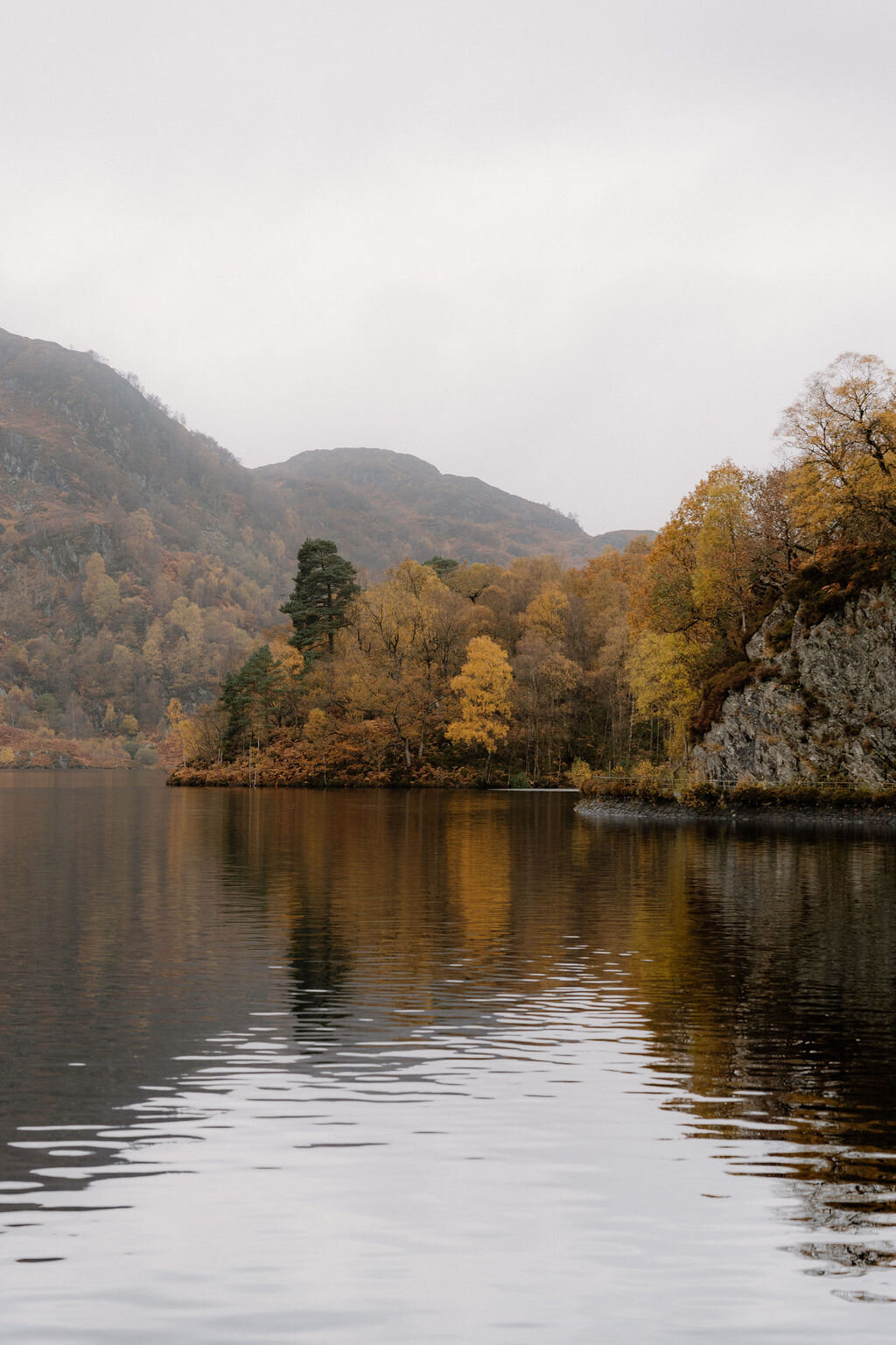 Autumn-coloured trees crowd the shore of a still loch near Wildwoodz, their reflection mirrored in the calm water beneath misty hills.