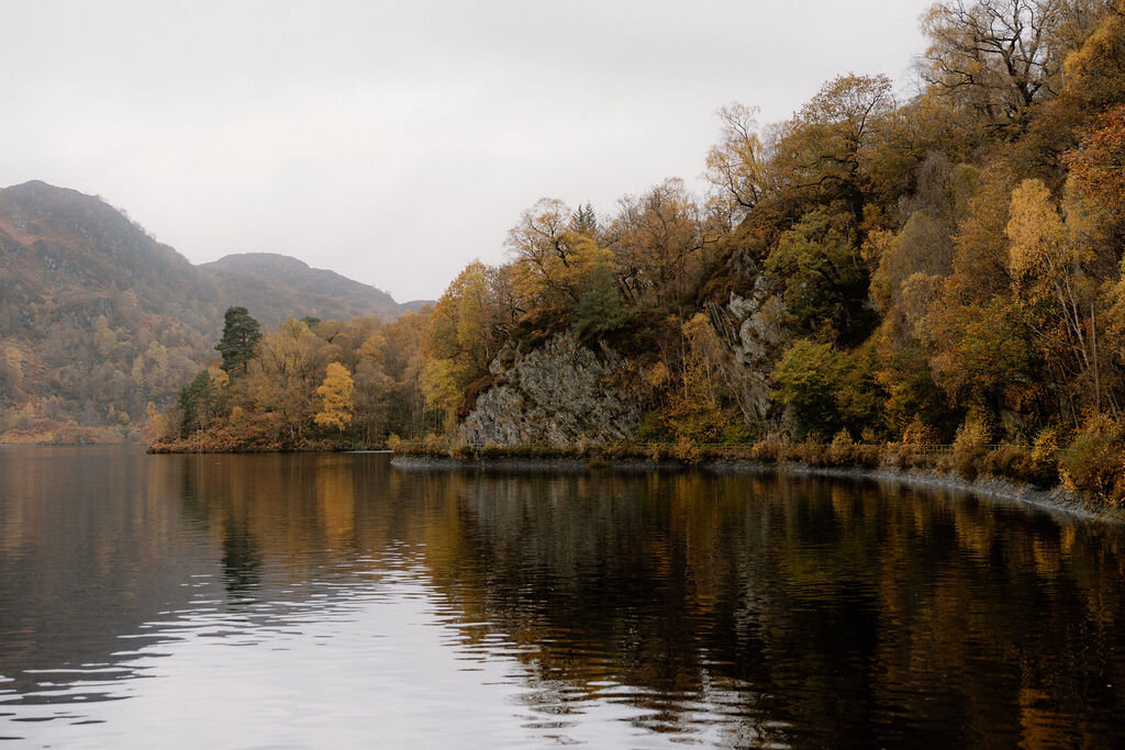 A rocky outcrop rises above autumn woodland reflected in a still loch near Wildwoodz, golden leaves lining the far shore.