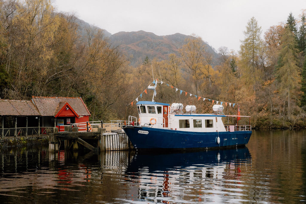 A tour boat moored with bunting sits beside a red-trimmed boathouse on a calm river near Wildwoodz, autumn trees and hills reflected in the water.