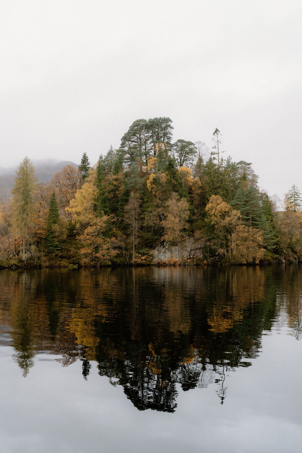 A small wooded island with autumn-coloured trees is mirrored in the still water of a loch near Wildwoodz.