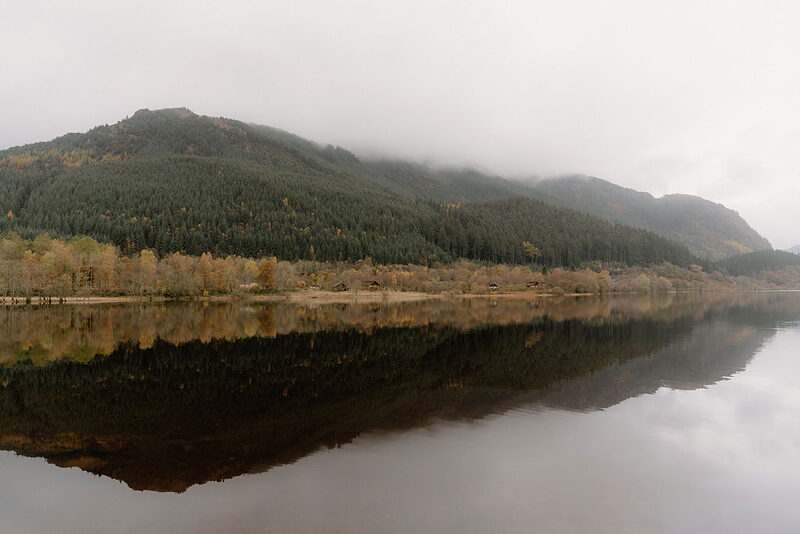 Forested hills reflected in a still loch at Wildwoodz beneath low autumn cloud.