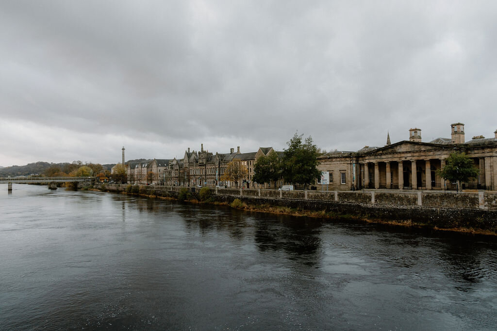 A wide river bordered by autumn trees, a bridge and a grand columned building on its far bank.