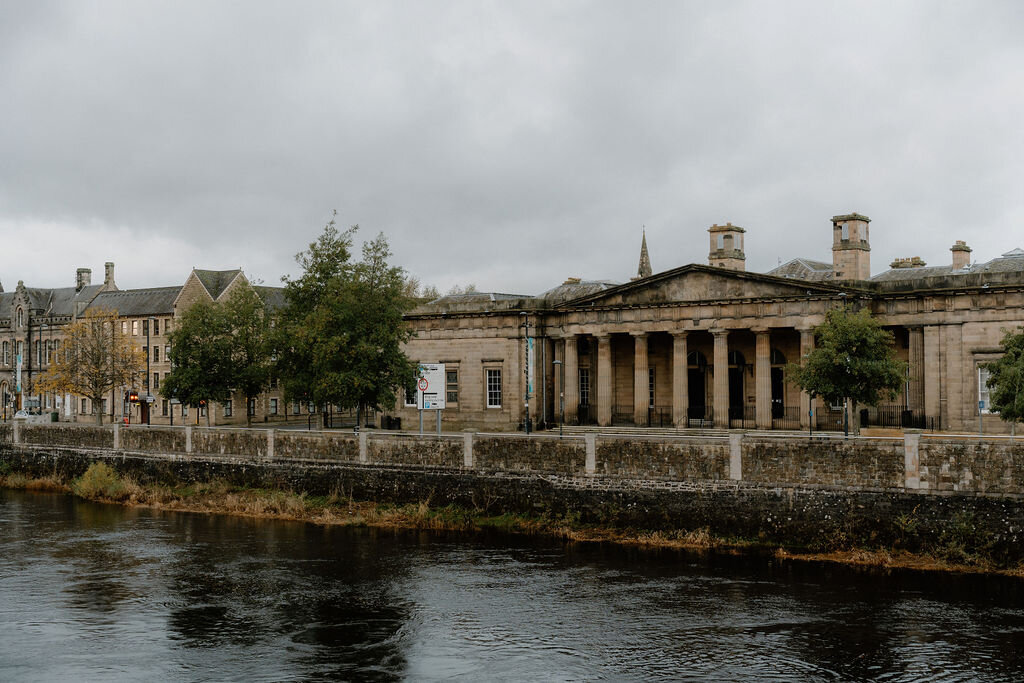 A river bordered by trees, a grand columned building standing on its bank.