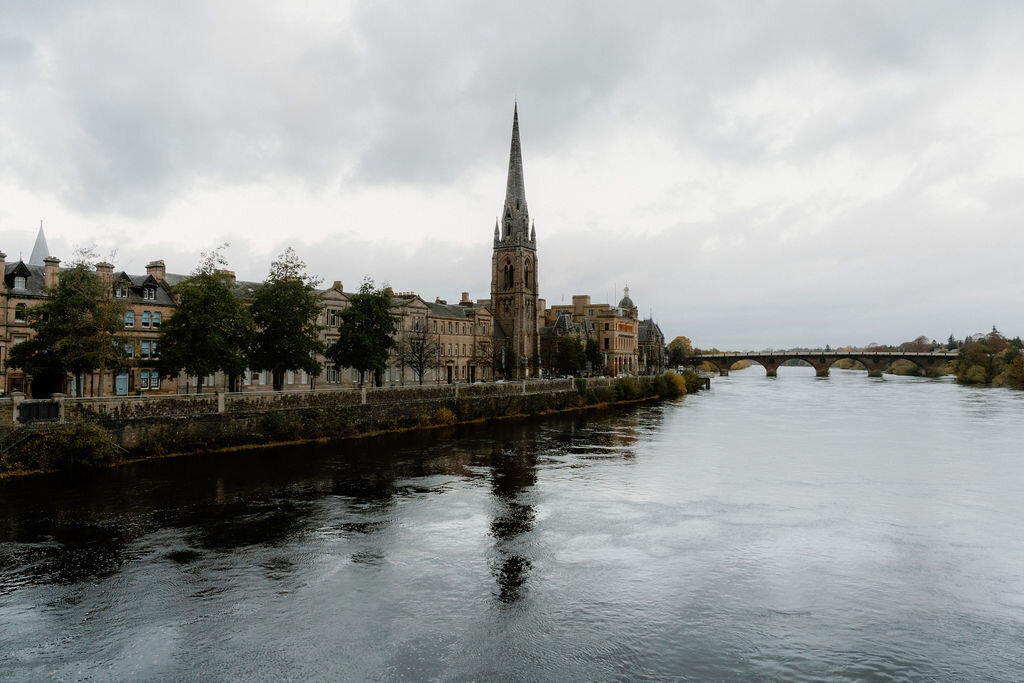 A river bordered by townhouses, a church spire and a bridge rising along its bank.