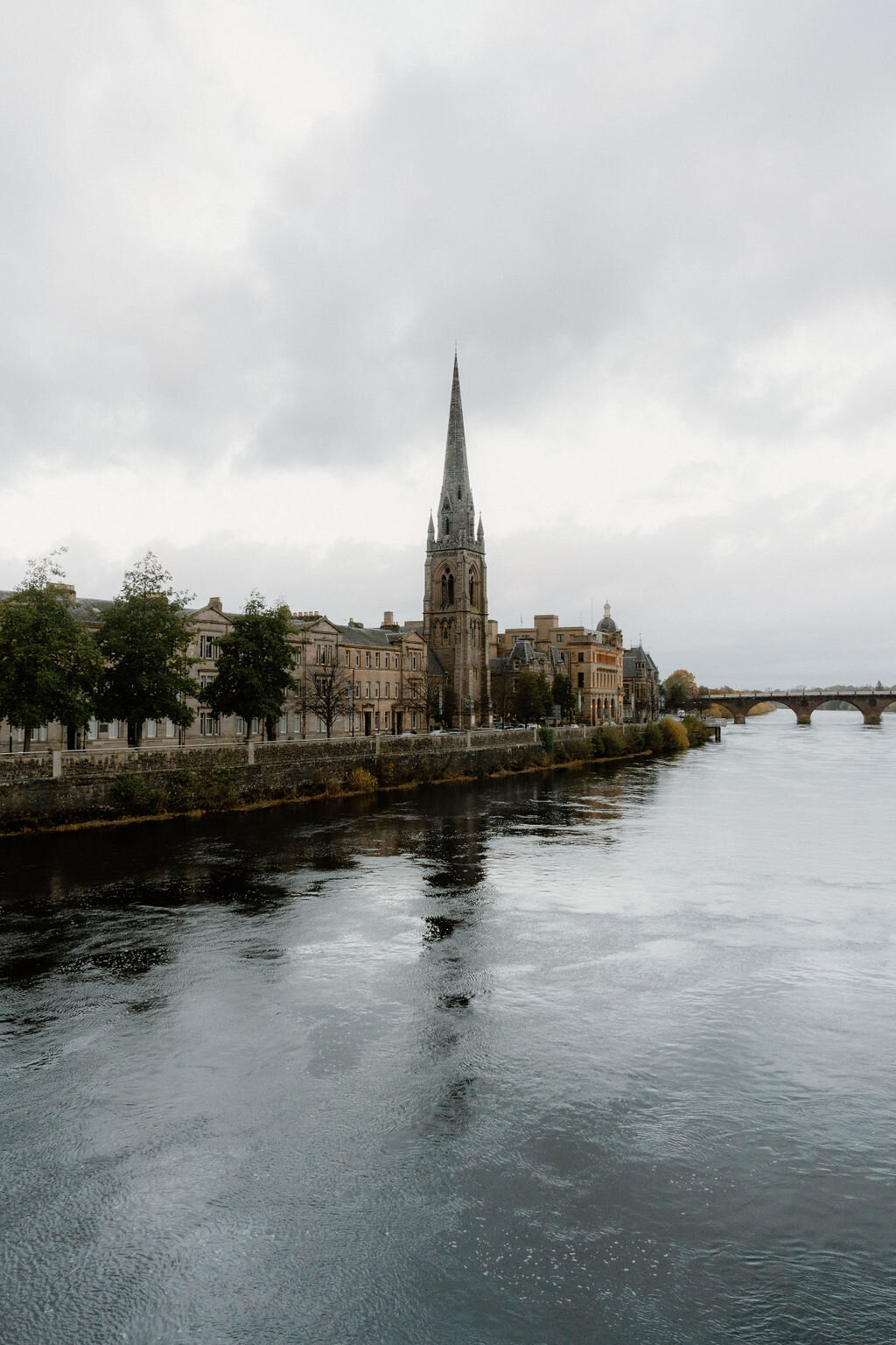 A river bordered by townhouses, a church spire and a bridge rising along its bank.