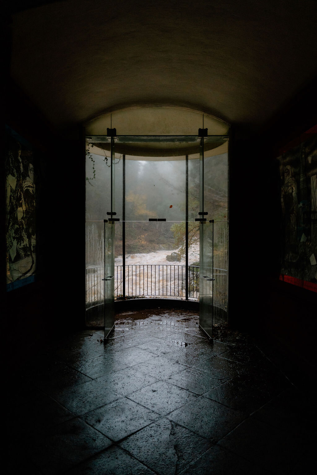 Glass doors open from a dim passage onto a misty river gorge, framed artworks lining the walls on either side.