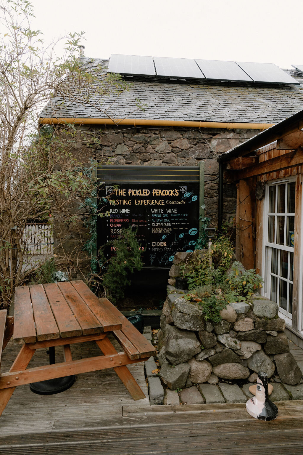 A chalkboard sign for The Pickled Peacock's tasting experience hangs on a stone wall beside a picnic table.