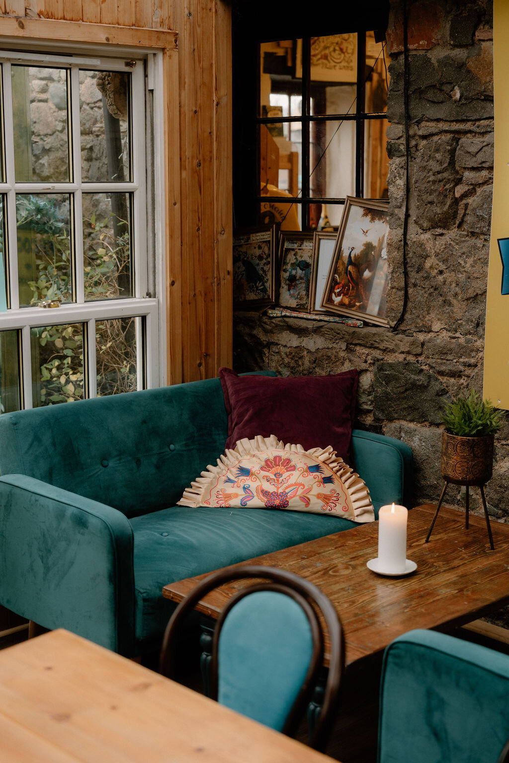 A teal velvet sofa with an embroidered cushion sits beside a stone wall and framed pictures inside The Pickled Peacock.