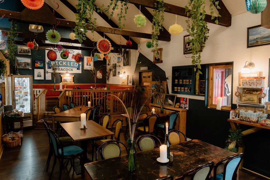 The Pickled Peacock's own sign sits on a shelf beneath hanging paper lanterns and trailing plants.