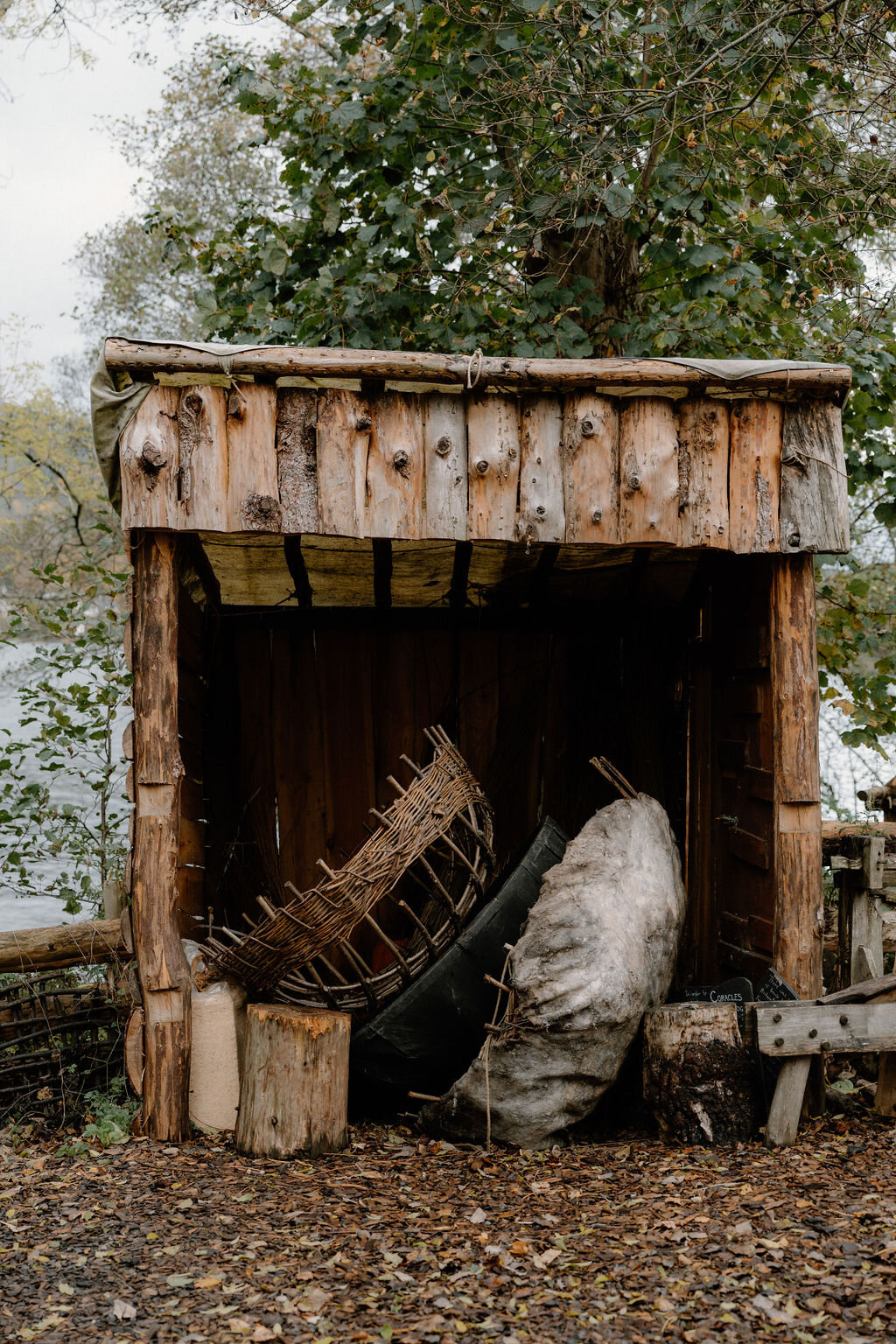 A woven wooden shelter holds coiled ropes and canvas bundles beside a riverbank.