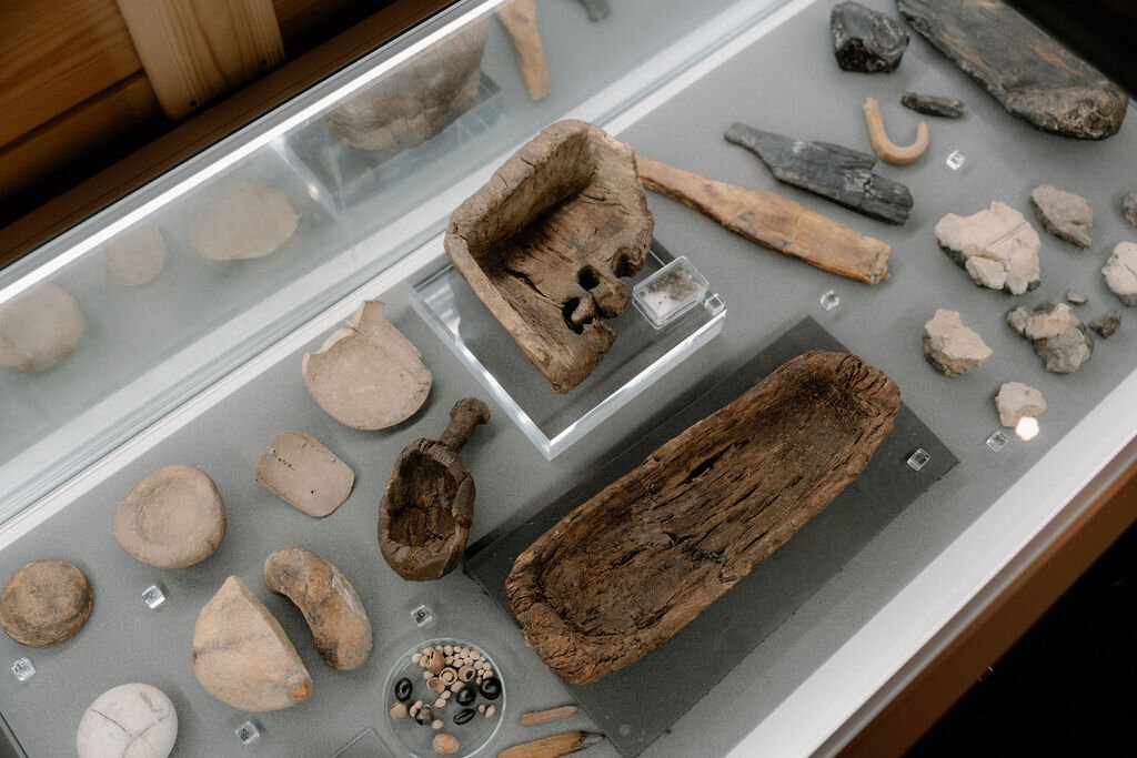 A museum case displays worked stones, a bone tool and a rusted cannonball on a pale surface.