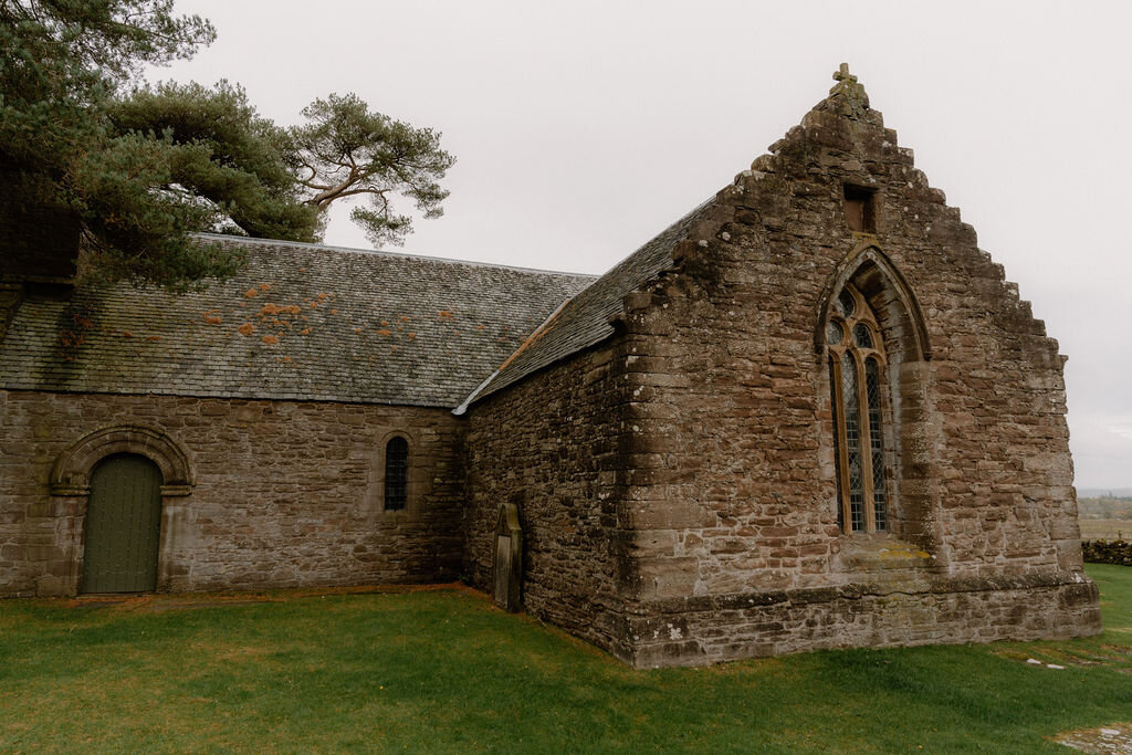A stone church building with buttressed gables stands beside a grassy churchyard.