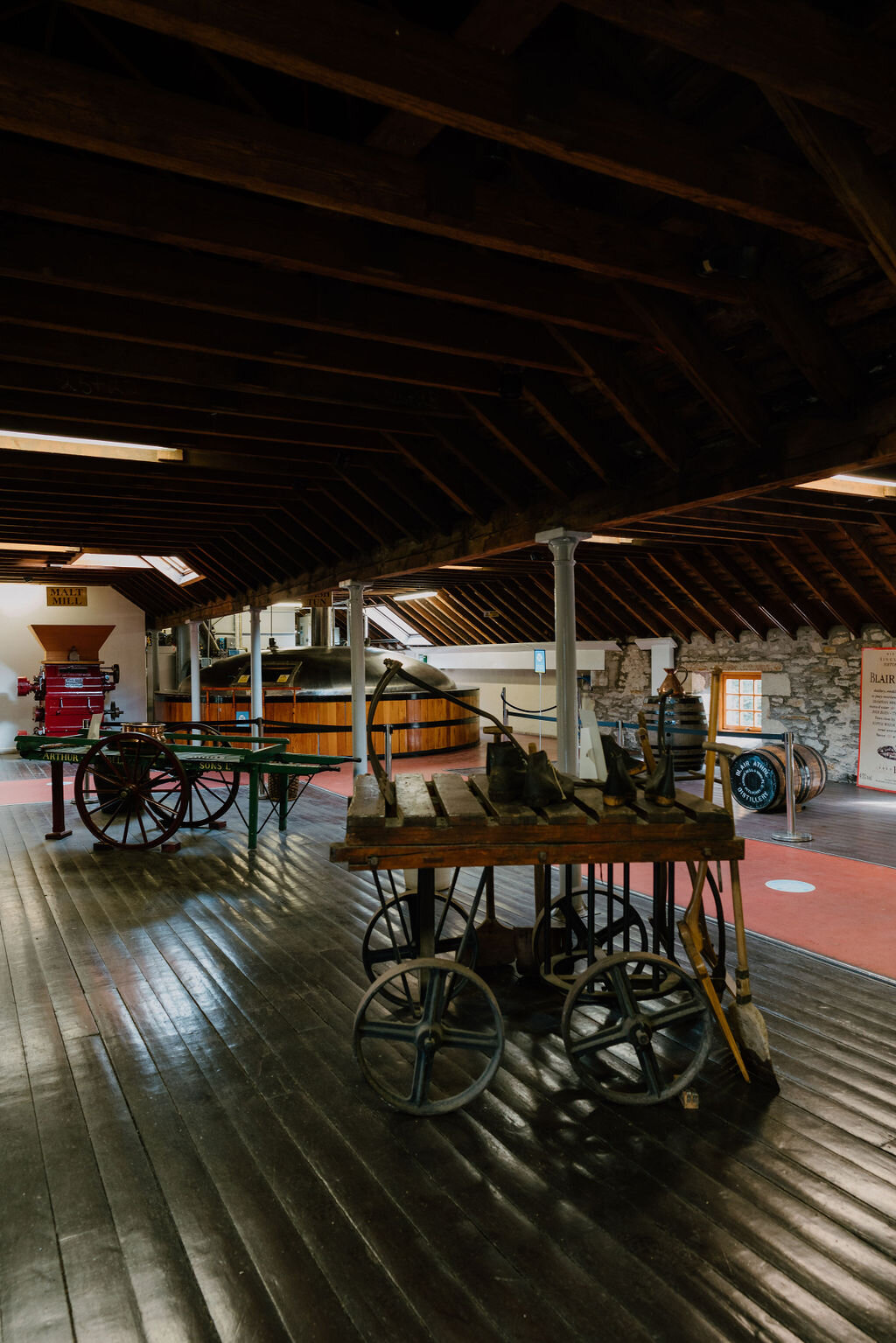 Antique wheeled carts and a red malt mill stand in a timber-roofed hall beside a copper mash tun at a Perthshire distillery.