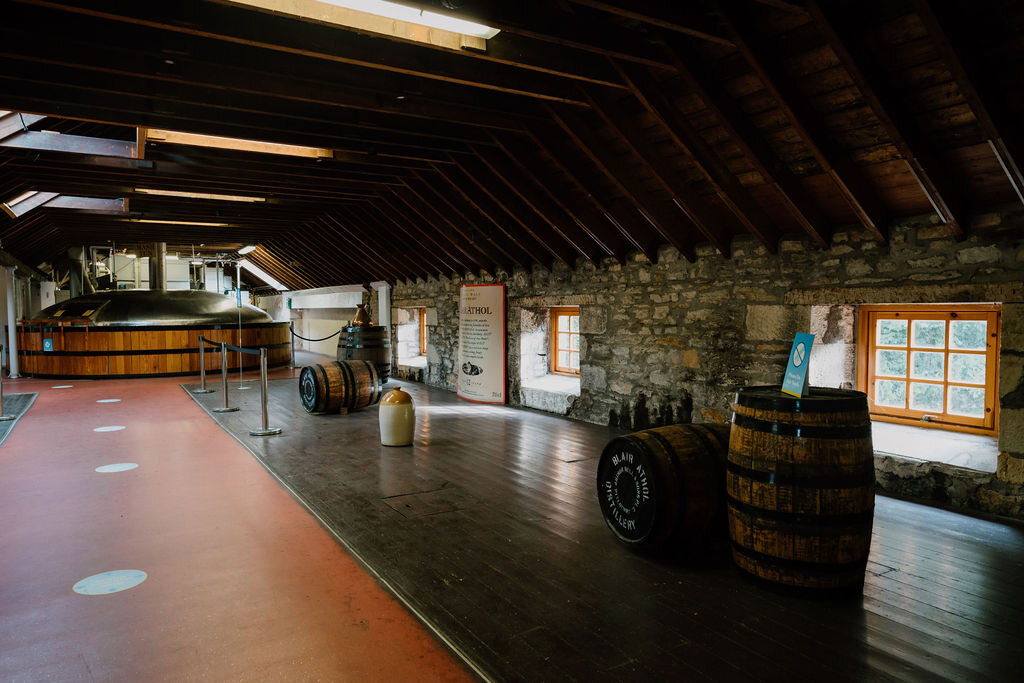 Whisky casks and an information stand sit in a timber-roofed exhibition hall beside a copper mash tun at a Perthshire distillery.