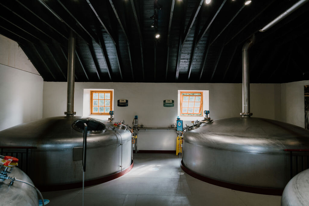 Two large steel mash tuns stand beneath a black timber roof, lit by small windows, at a Perthshire distillery.