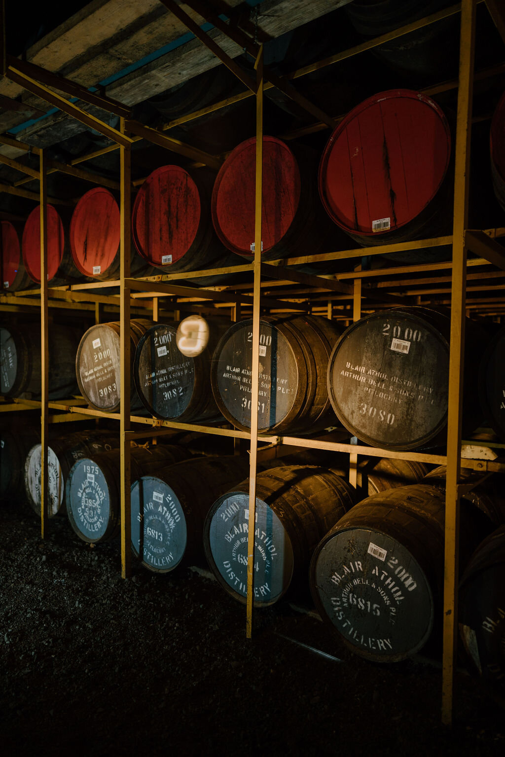 Casks stamped 'BLAIR ATHOL DISTILLERY' are stacked on racks, some with red-painted heads, in a dim warehouse.