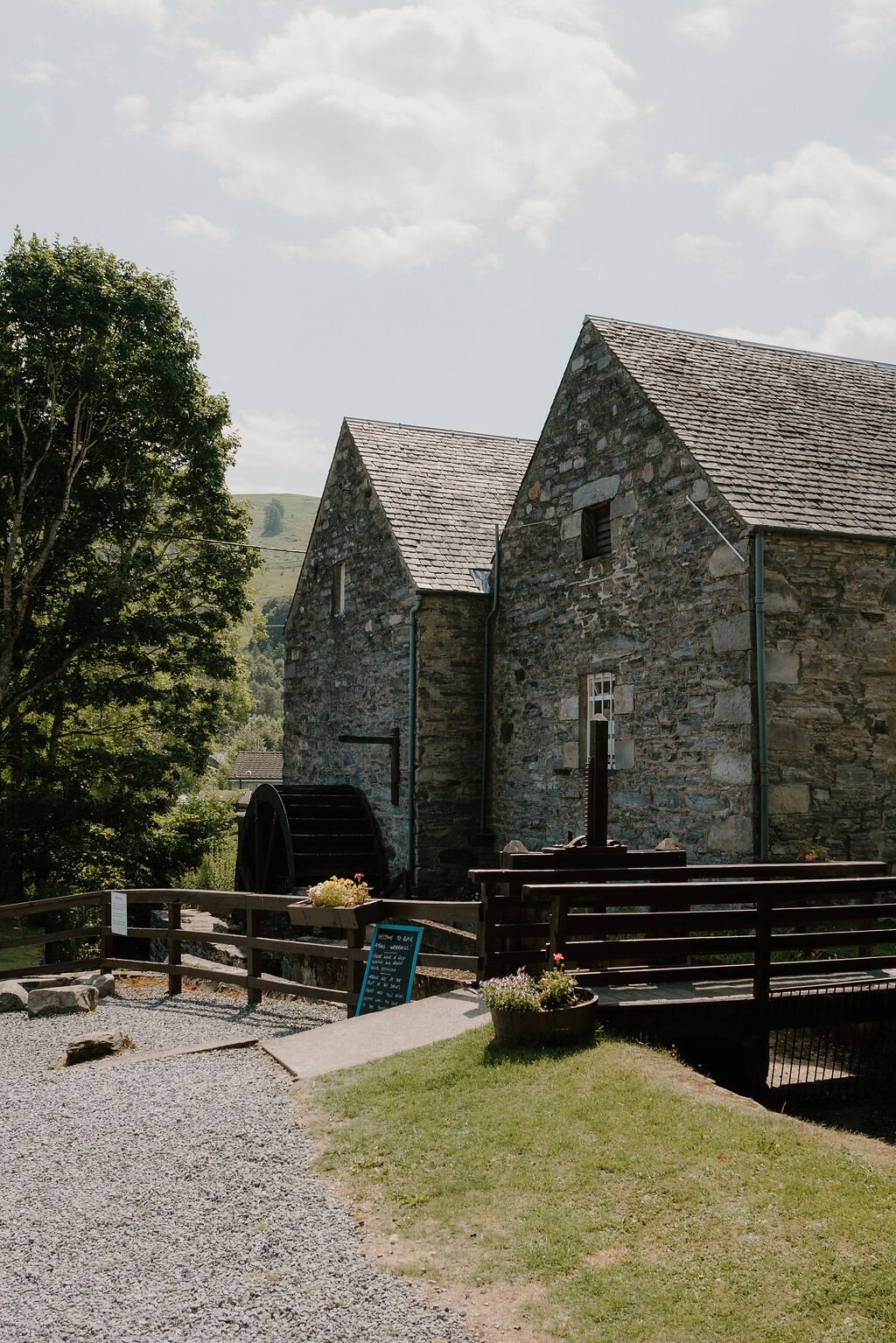 A stone watermill with a waterwheel and flower-filled planters stands beside a wooden walkway.