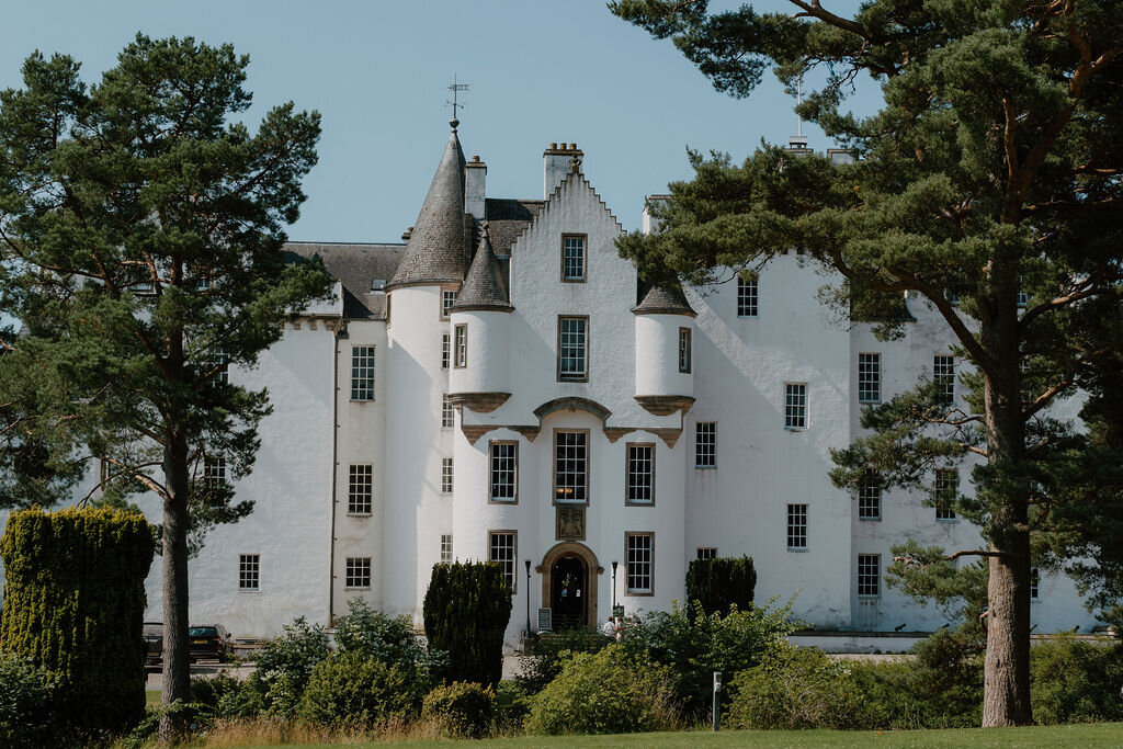 Blair Castle's white harled facade and conical turrets stand behind a screen of pine trees.