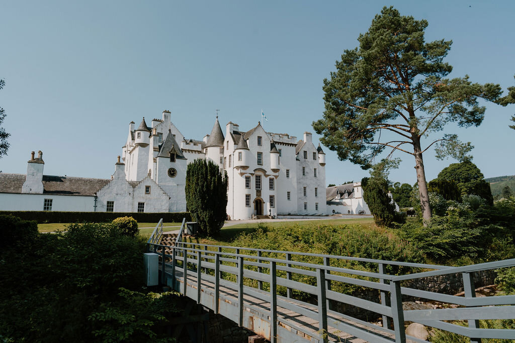 A wooden footbridge leads toward Blair Castle's white facade, framed by a mature pine tree.