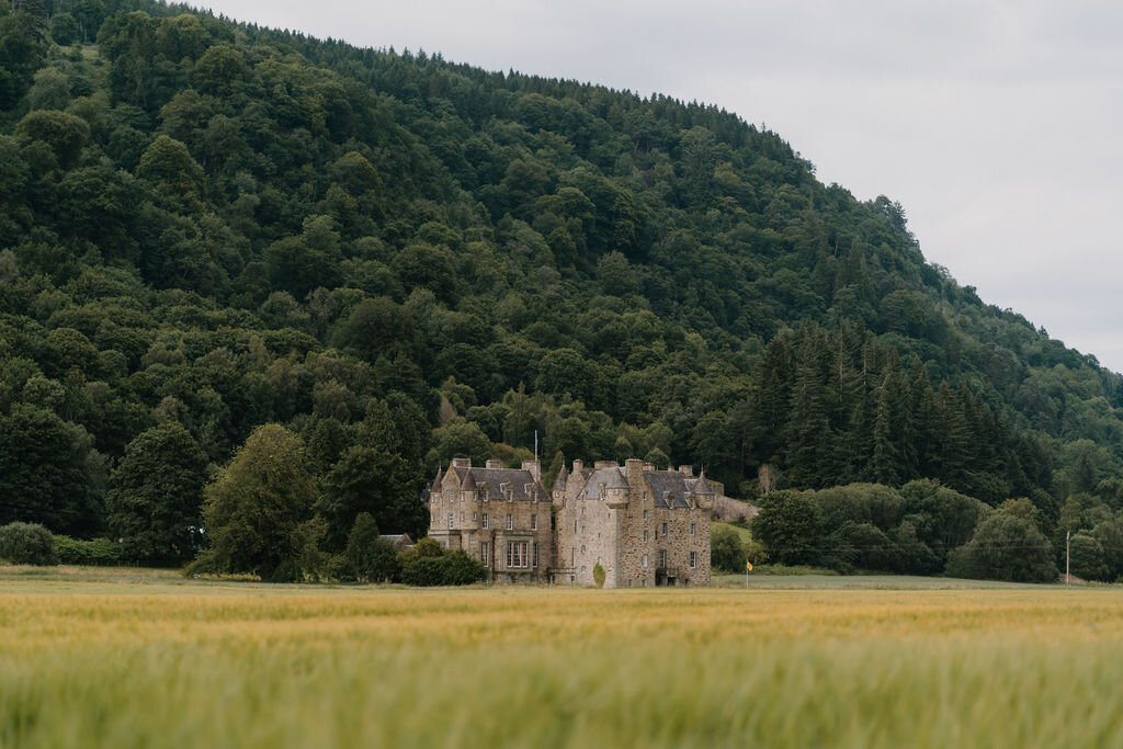 A distant turreted castle stands before a densely forested hillside beyond a barley field.