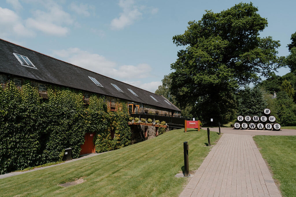 Ivy climbs the gabled stone wall of Dewar's Aberfeldy distillery beside a paved path.