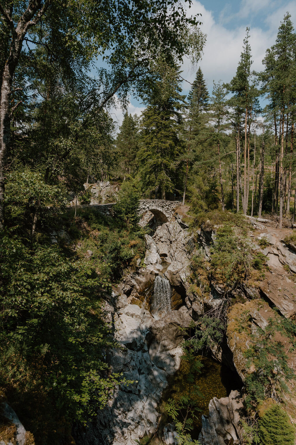 An old stone packhorse bridge arches over a rocky gorge and waterfall in a Perthshire forest.