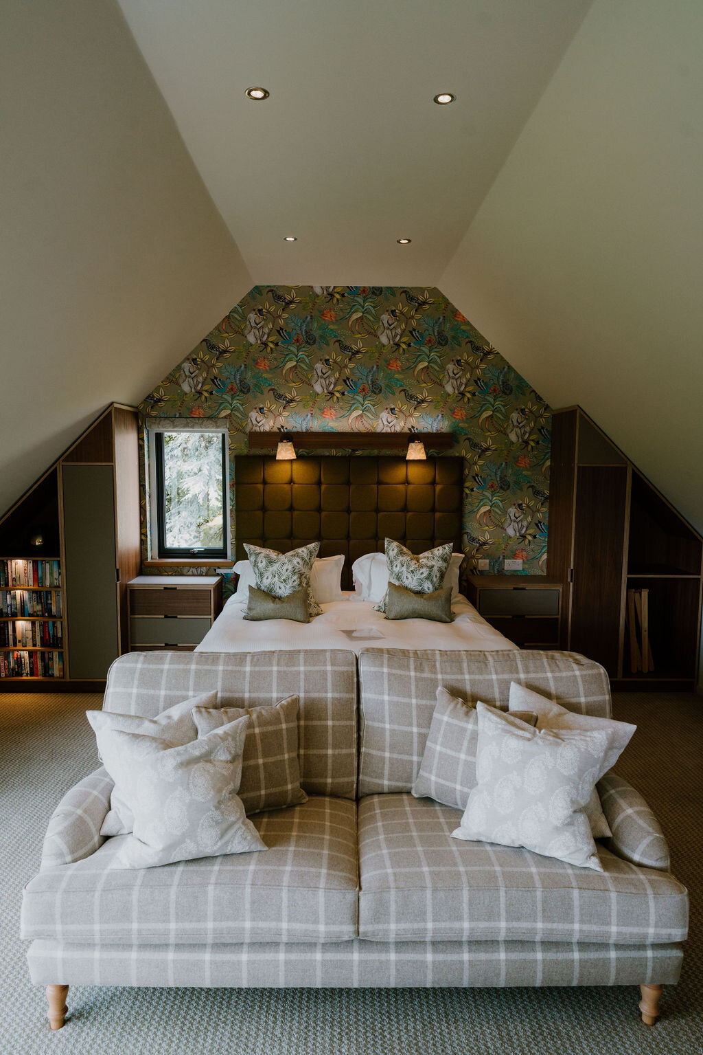 A bed with patterned headboard wallpaper sits beneath a sloped ceiling, a checked sofa at its foot.