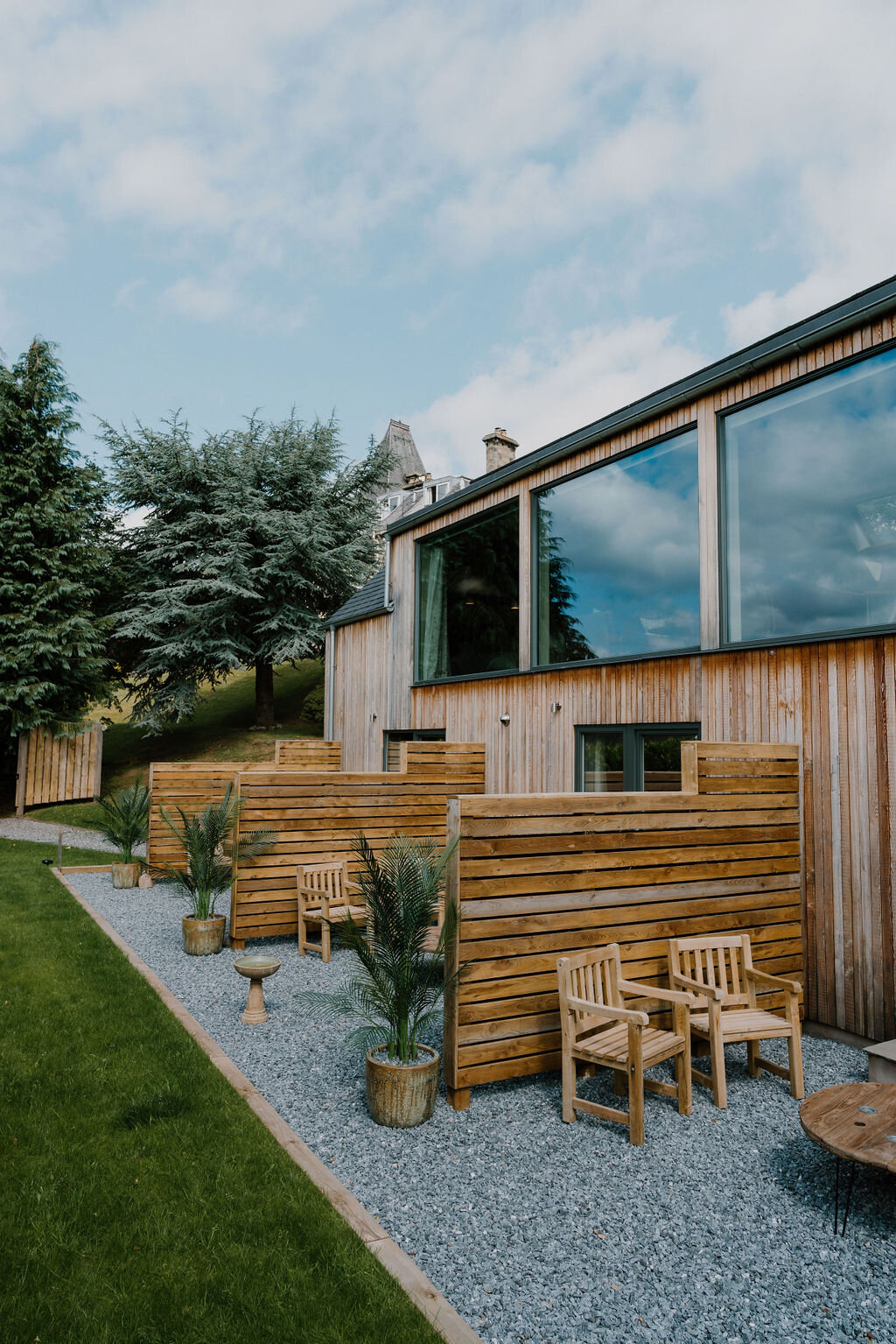Wooden slatted screens and rattan chairs furnish a gravel terrace beside a timber-clad building, a castle tower visible on the hillside beyond.
