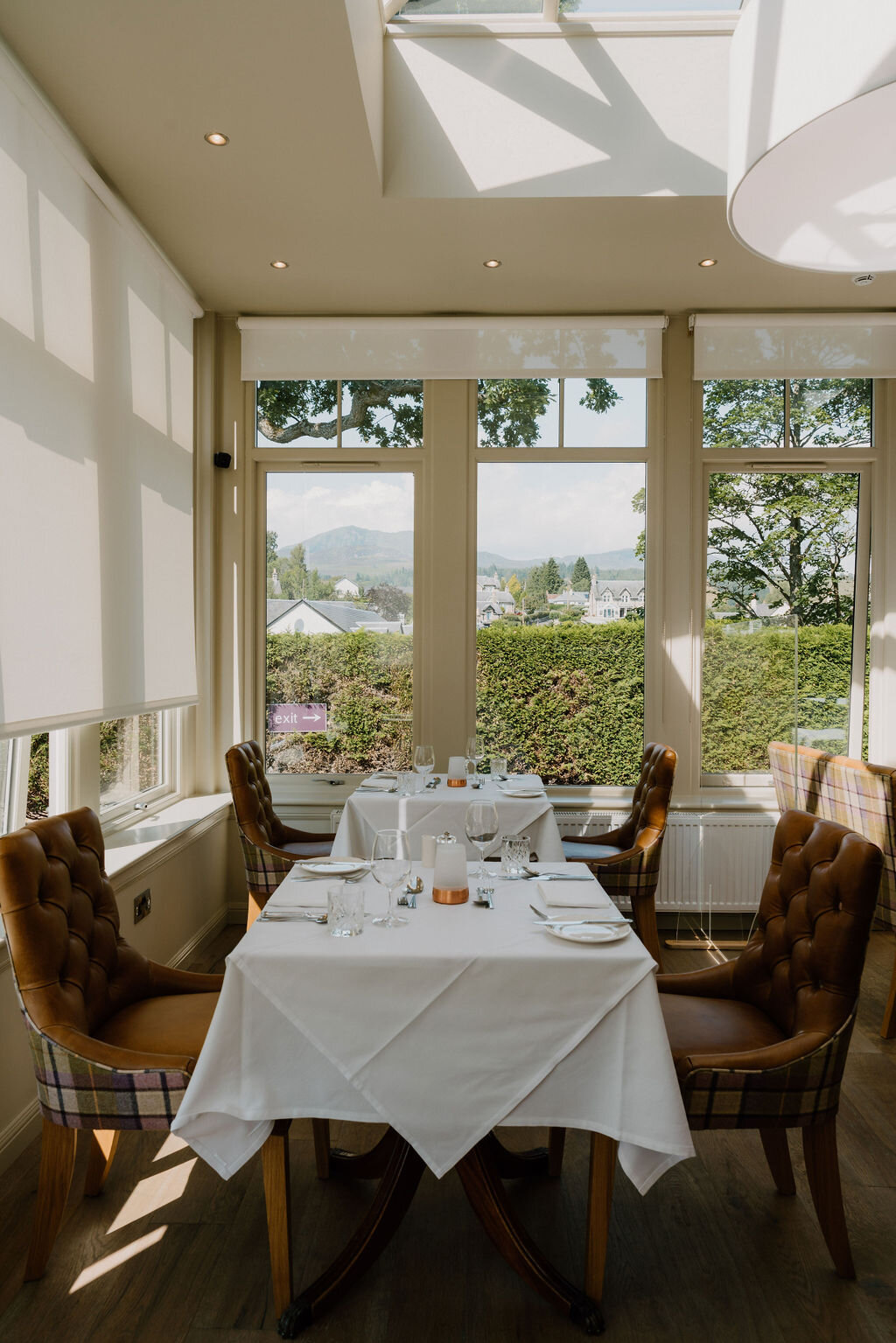 A table set for four sits beneath a glass roof lantern in a conservatory dining room overlooking a village.