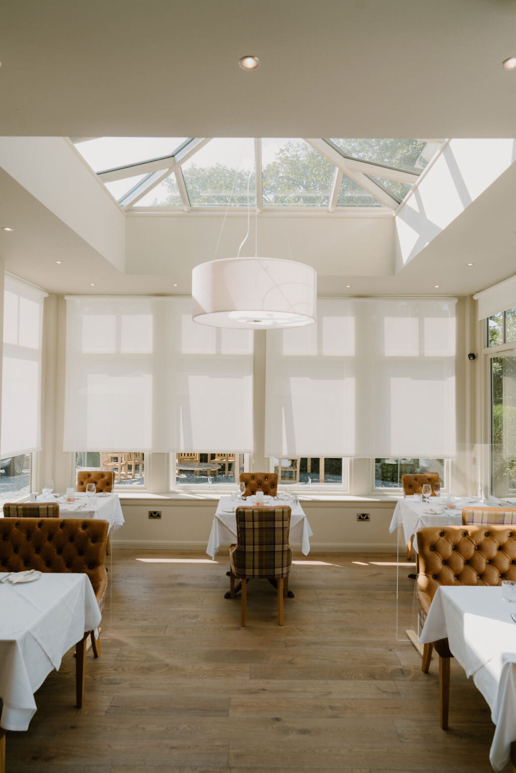 Tartan-upholstered chairs surround white-clothed tables beneath a glass atrium roof in a conservatory dining room.