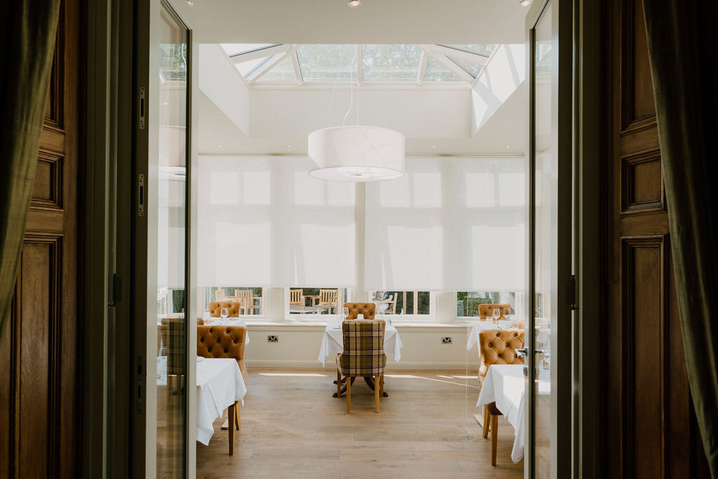 Open double doors reveal a sunlit conservatory dining room with tartan chairs and a pendant light.