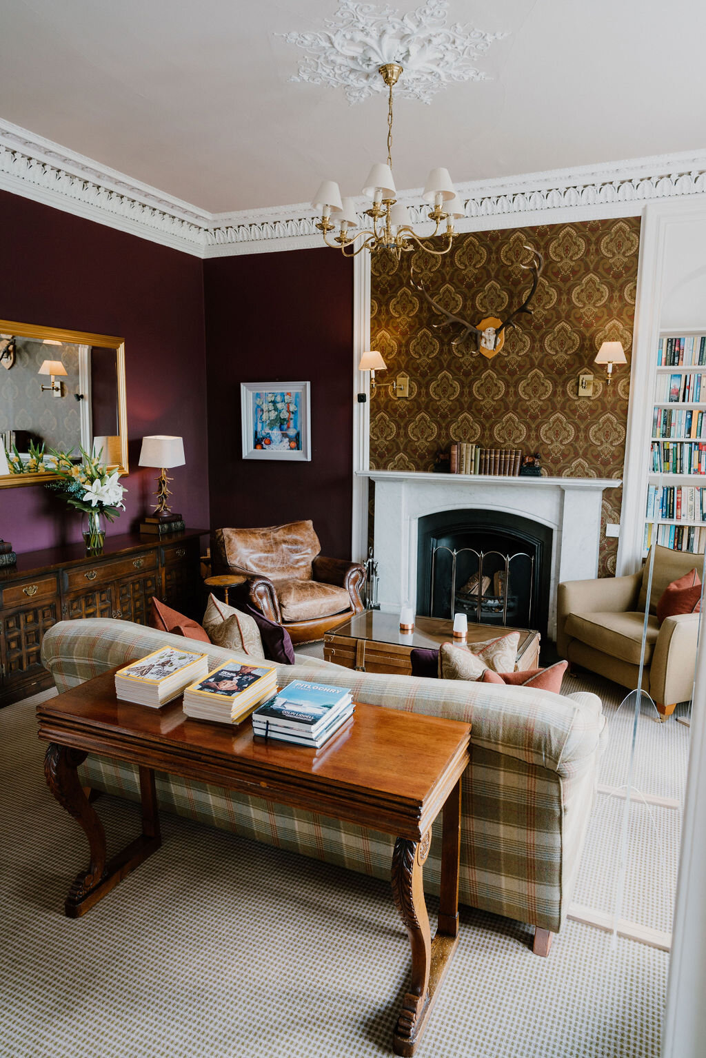 A burgundy drawing room has a marble fireplace, antler mount and tartan sofa beneath an ornate chandelier.