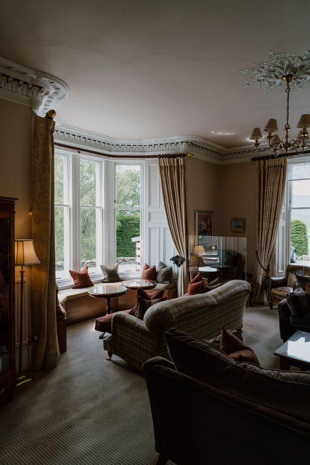 A bay-window drawing room with tartan sofas and a chandelier looks out over a garden.