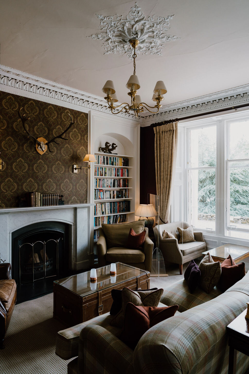 A drawing room with patterned wallpaper, a mounted stag's head and a bookcase surrounds a marble fireplace.