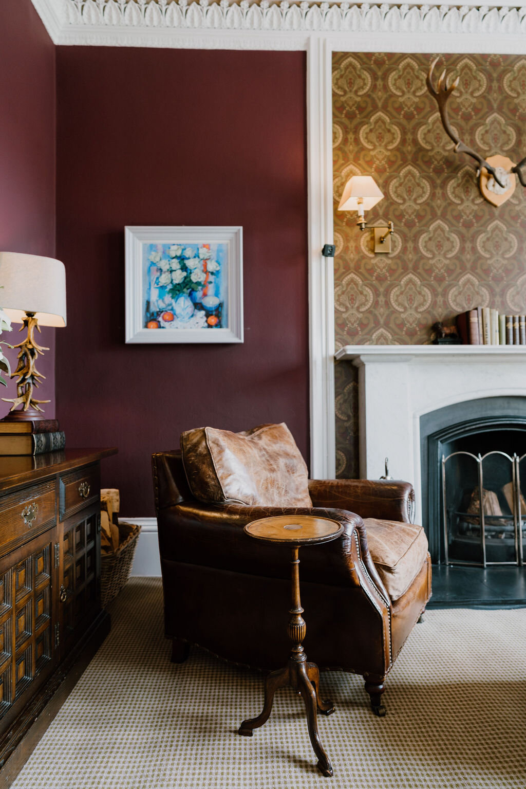 A leather armchair sits beside a fireplace beneath a mounted stag's head in a burgundy-walled room.