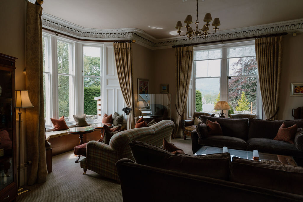 A bay-window drawing room with sofas and armchairs overlooks a garden through tall curtained windows.