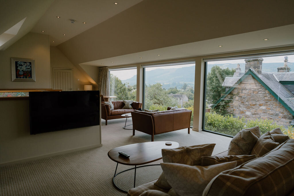 Leather armchairs and a corner sofa face a large window overlooking hills and a church spire.