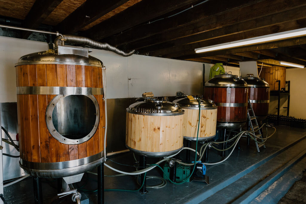 A row of small wooden-clad fermentation tanks connected by hoses fills a low-ceilinged brewery room.