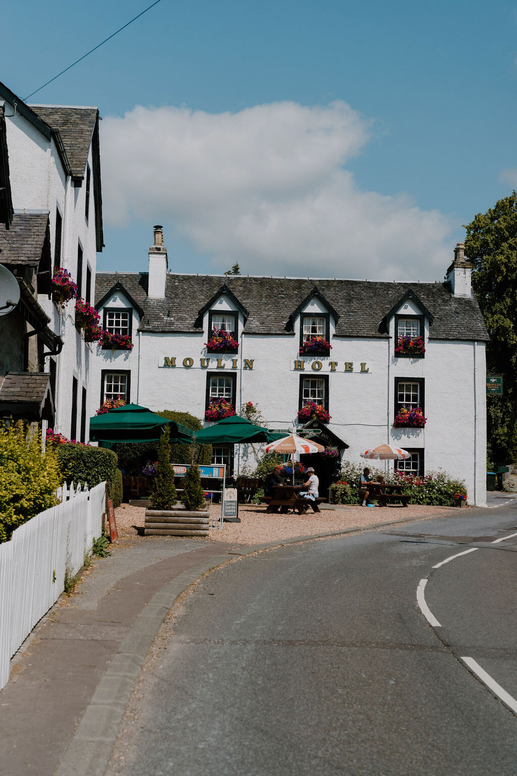 The Moulin Hotel's white harled facade is lettered above outdoor tables shaded by green umbrellas.