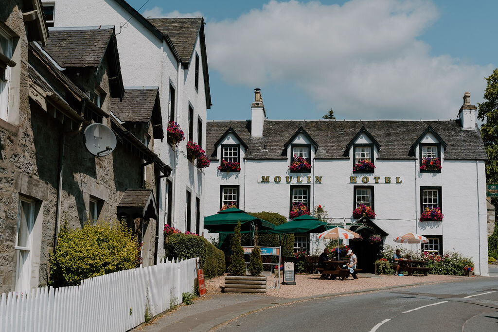 The Moulin Hotel's white facade with hanging flower baskets overlooks outdoor tables shaded by umbrellas.