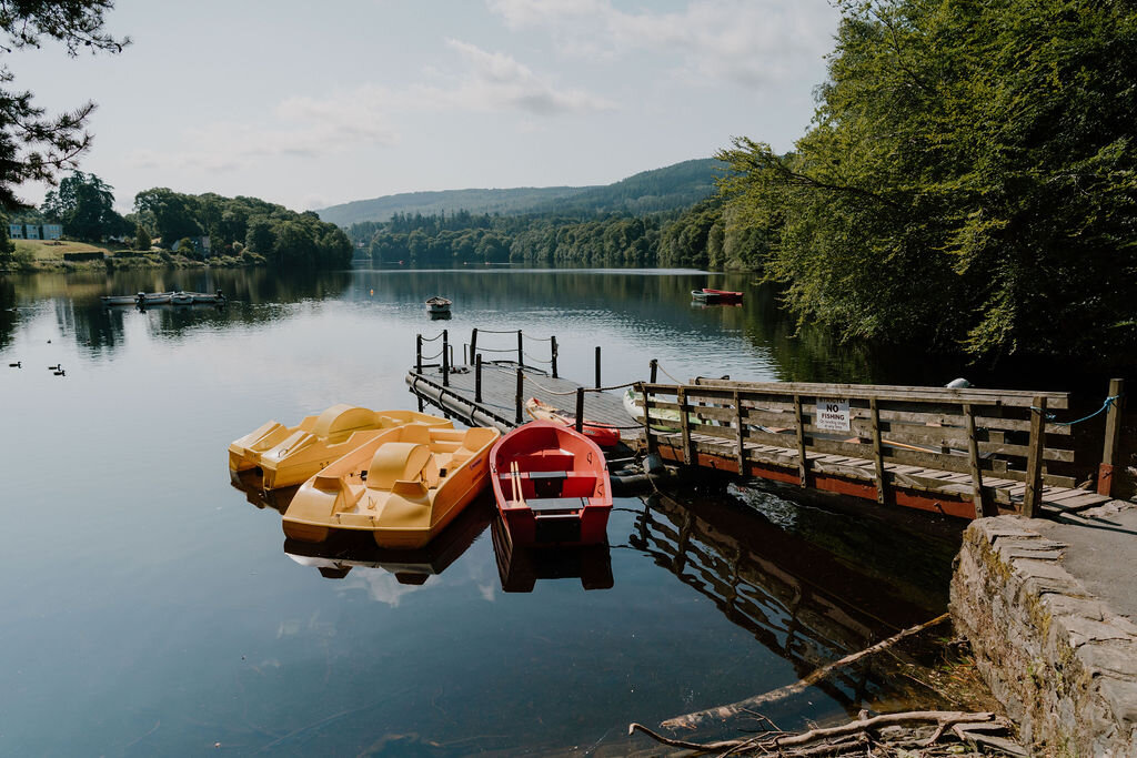 Yellow and red pedalos are moored at a wooden jetty on a calm loch beneath wooded hills.