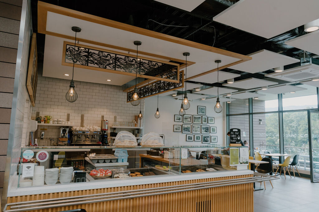 A cafe counter with pastries and cakes sits beneath pendant lights and an ornate metal canopy.