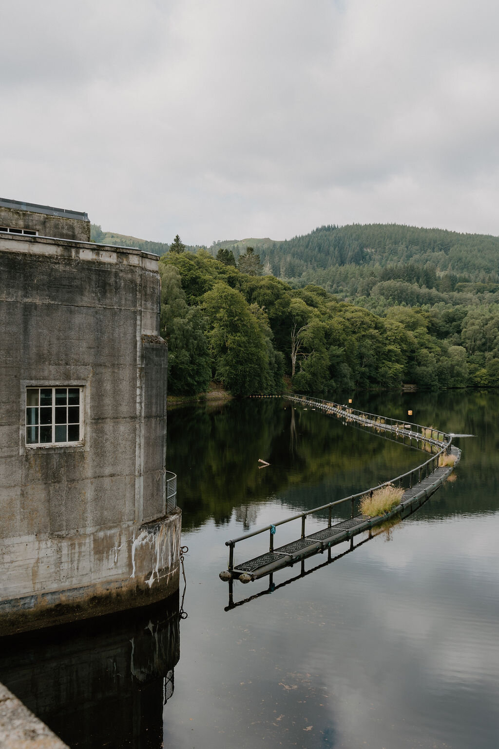 A footbridge crosses Loch Faskally beside a concrete dam building surrounded by wooded hills.