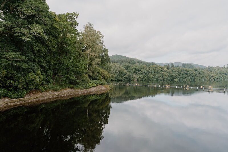 Wooded hills and a distant fish-pass walkway are mirrored in the calm surface of Loch Faskally.