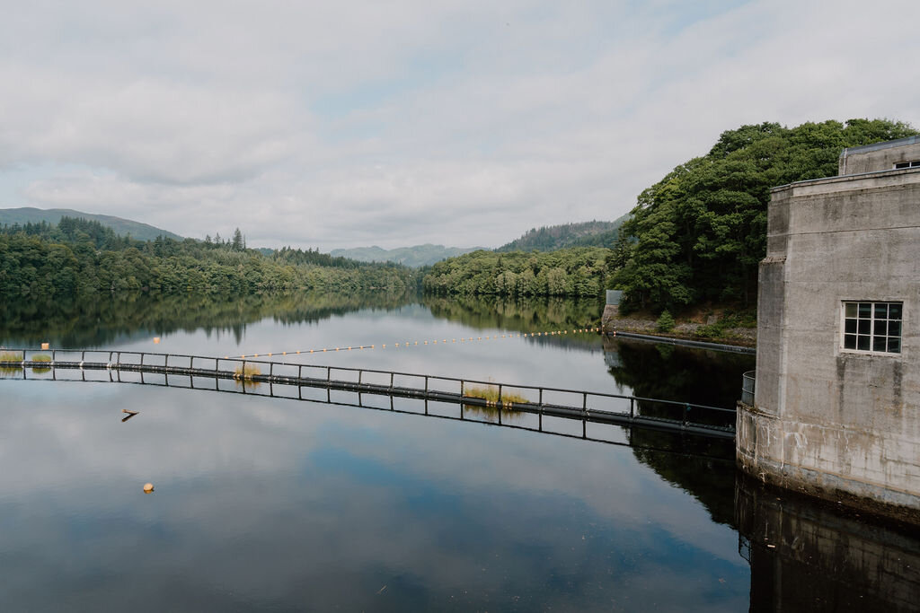 A fish-pass walkway crosses Loch Faskally beside the dam building, mirrored in still water.