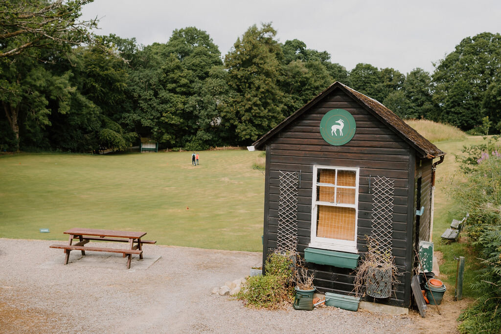 A dark timber hut with a crest sign stands beside a mown putting green and picnic table.