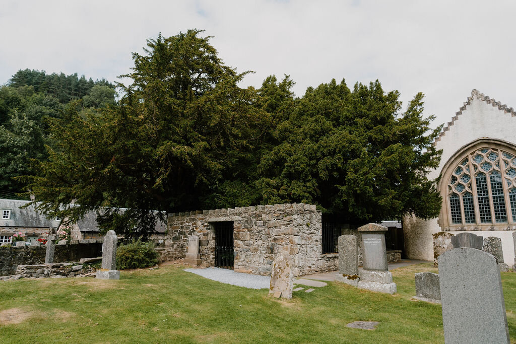 Gravestones stand before a stone churchyard wall and a Gothic church window shaded by ancient yew trees.