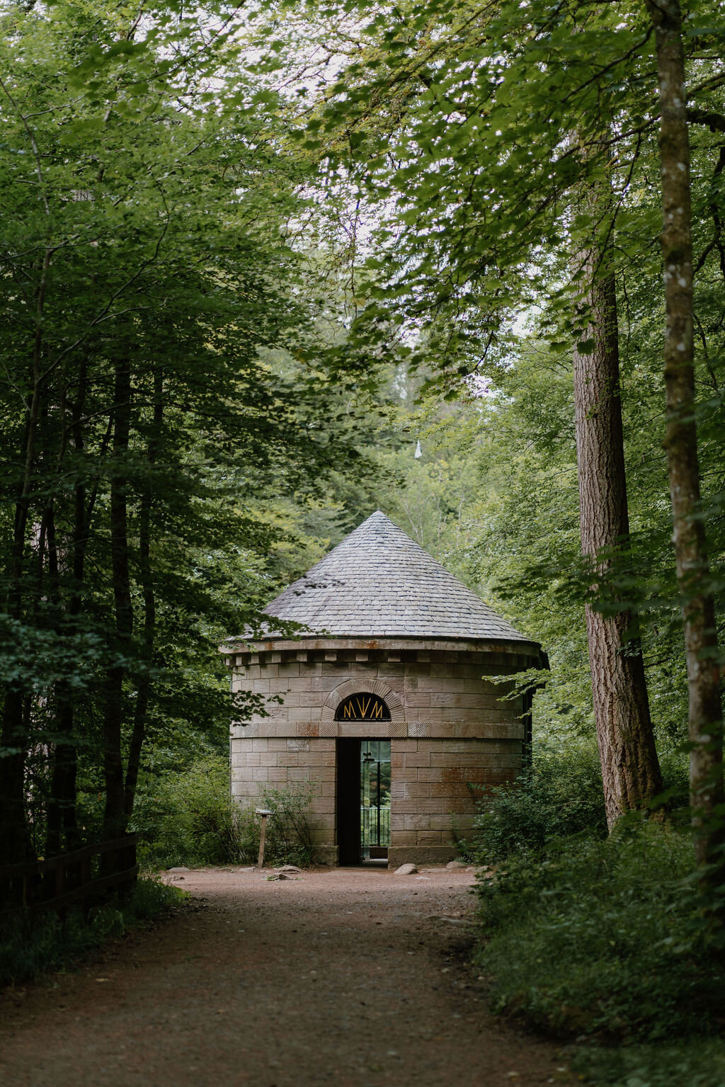 A small round stone folly with a conical slate roof and arched doorway stands among trees in a Perthshire wood.